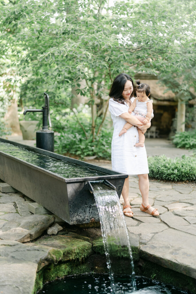 Mother and toddler daughter dressed in white linen dress pose near the water fountain at Winterthur Gardens. 