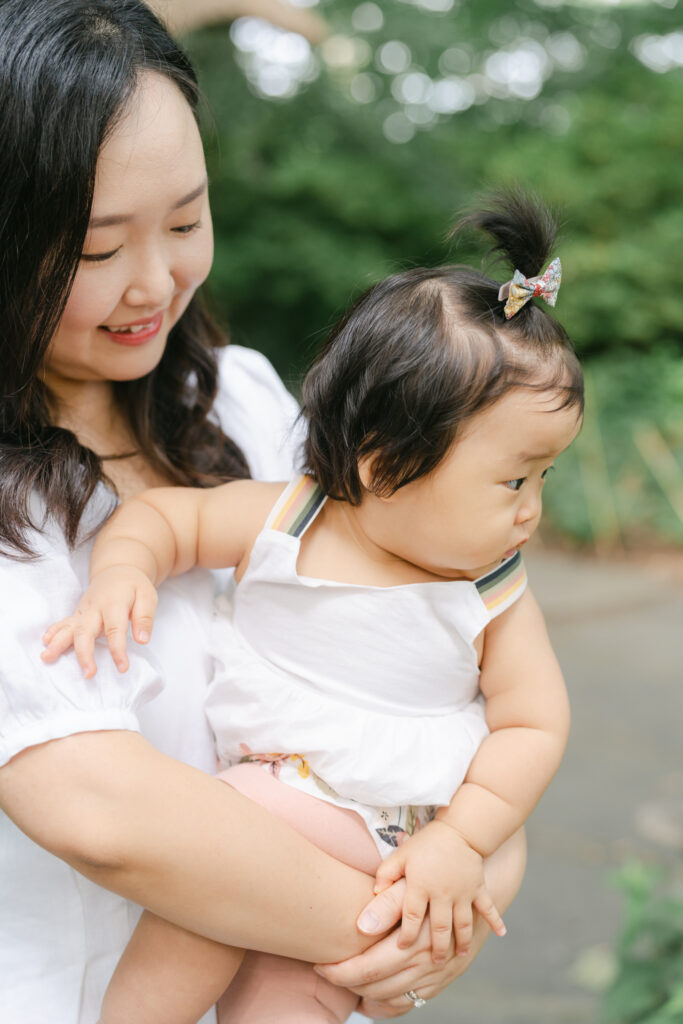 Dark-haired mother holds dark-haired baby with pigtail and white dress. 