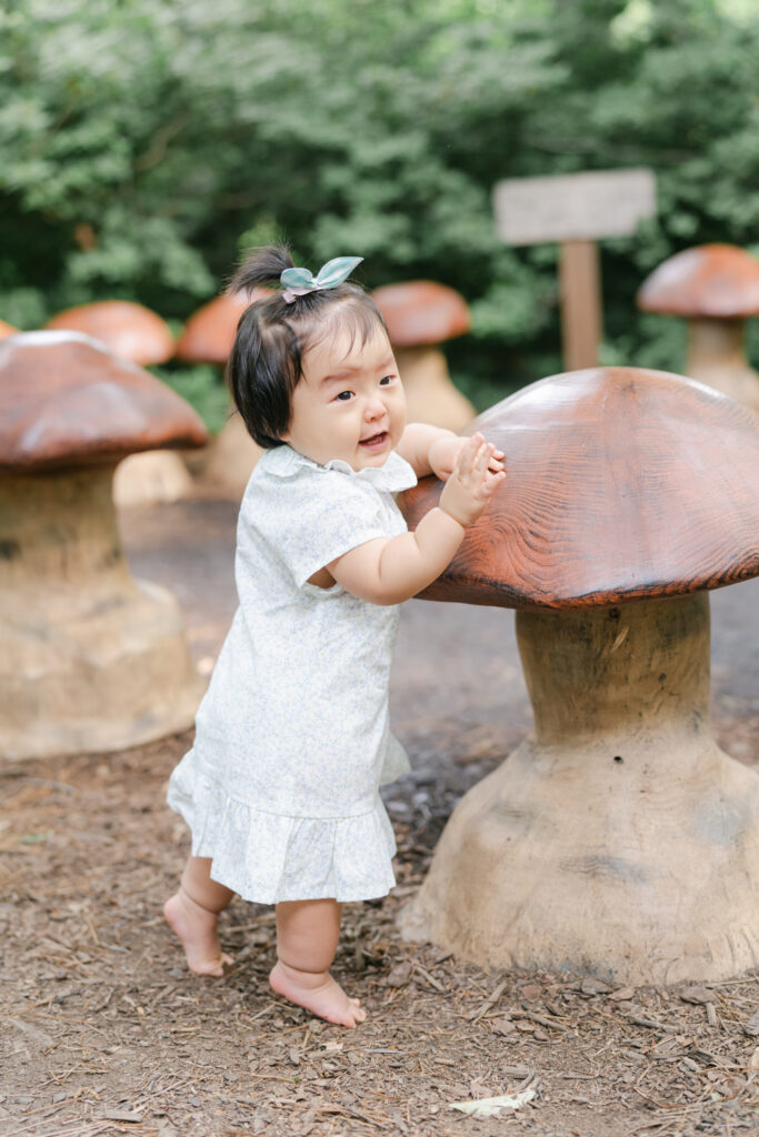 Toddler with dark hair in dress poses for a photo at the misting mushrooms in Winterthur Gardens in Wilmington DE
