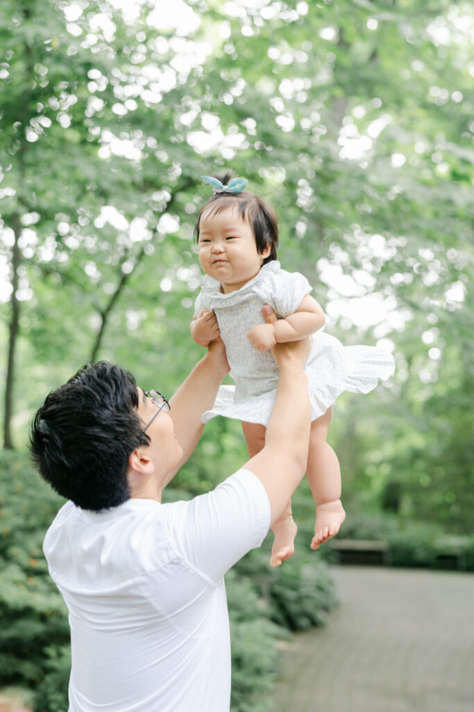 Father holds baby girl with pigtail and dress up in the air during family photos with AnneMarie Hamant