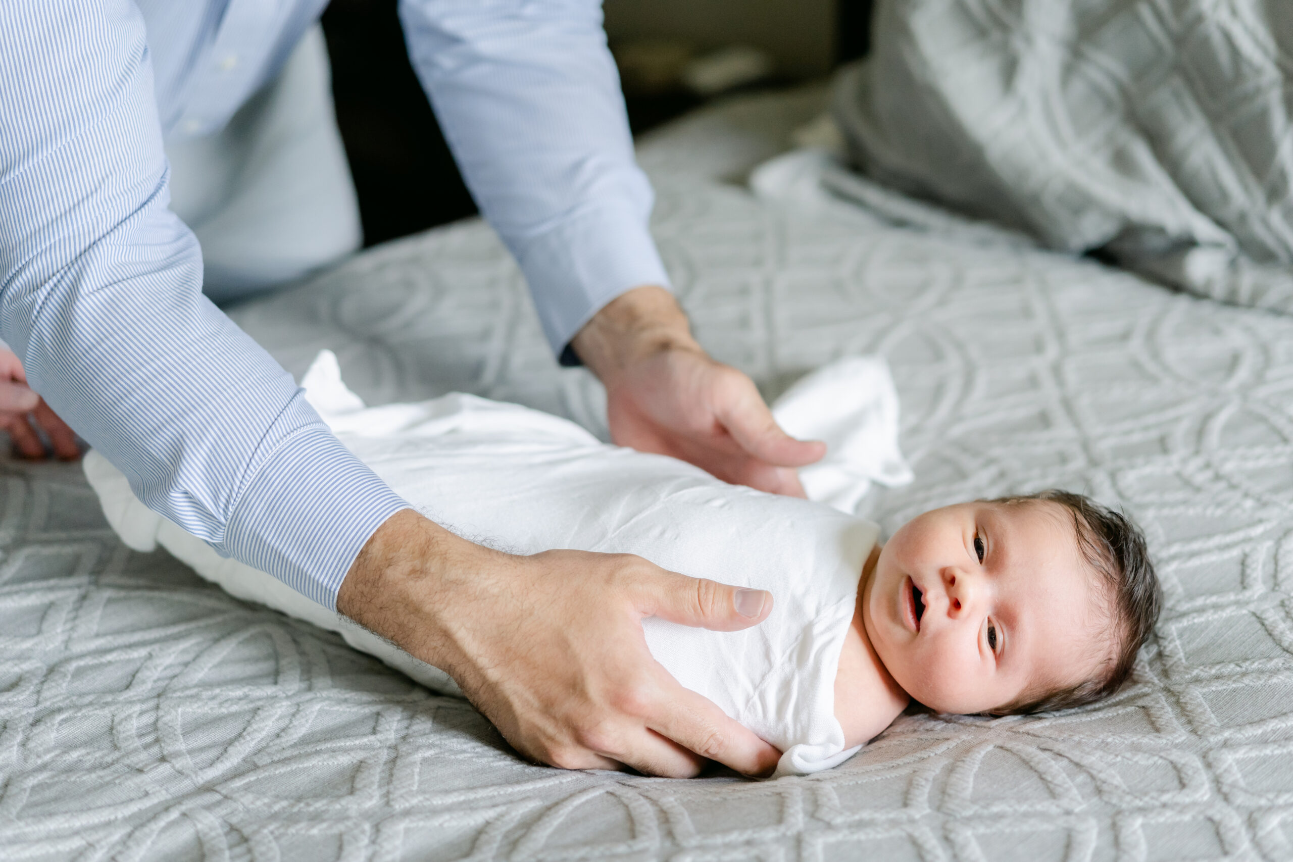 newborn wrapped in a white swaddle laying on a bed with dad's hands Philadelphia Baby Stores