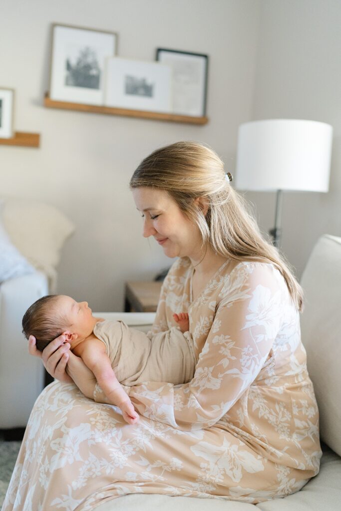 mom sitting on the couch in a neutral dress staring at newborn son