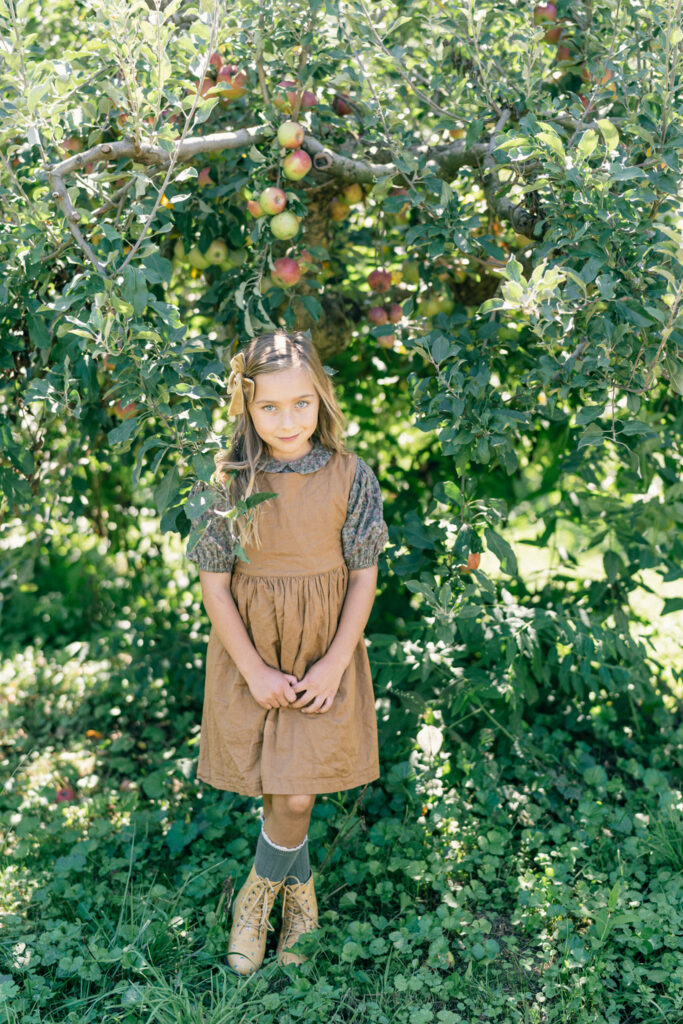 Girl in linen dress and yellow boots and stands in front of an apple tree while apple picking near Wilmington, DE
