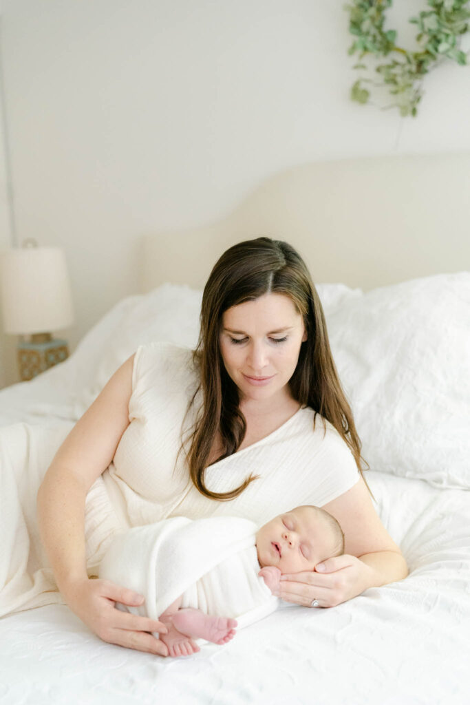 Mother in white dress cuddles her swaddled newborn baby in her Wilmington, DE home during a newborn session with AnneMarie Hamant