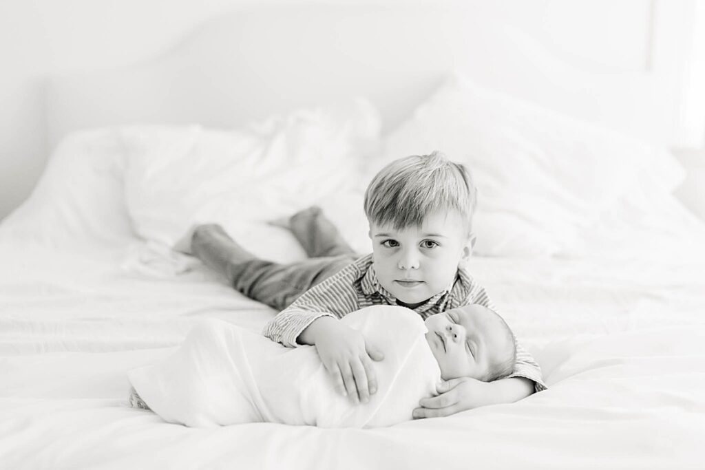 Black and white picture of big brother laying on the bed with baby brother sleeping by wilmington de newborn photographer AnneMarie Hamant