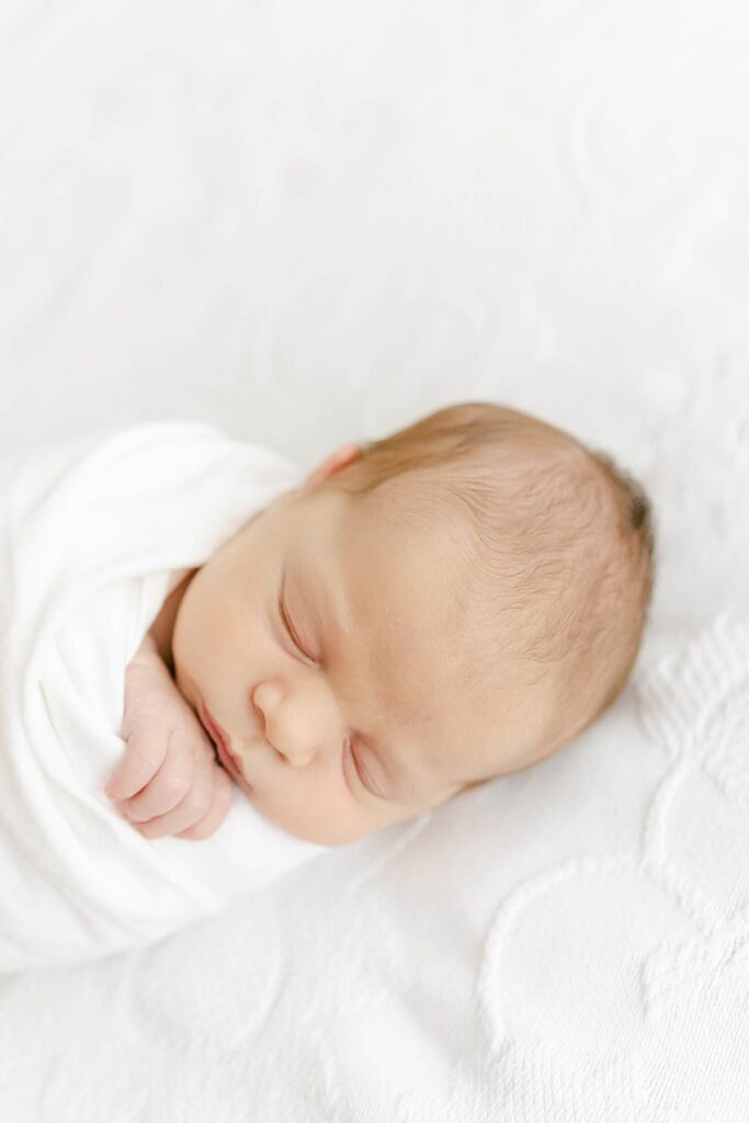 newborn boy swaddled in white wrap laying on a white bed comforter by wilmington de newborn photographer, AnneMarie Hamant