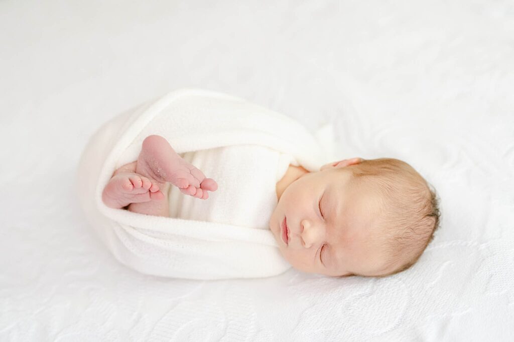newborn boy swaddled in white wrap laying on a white bed comforter by wilmington de newborn photographer, AnneMarie Hamant