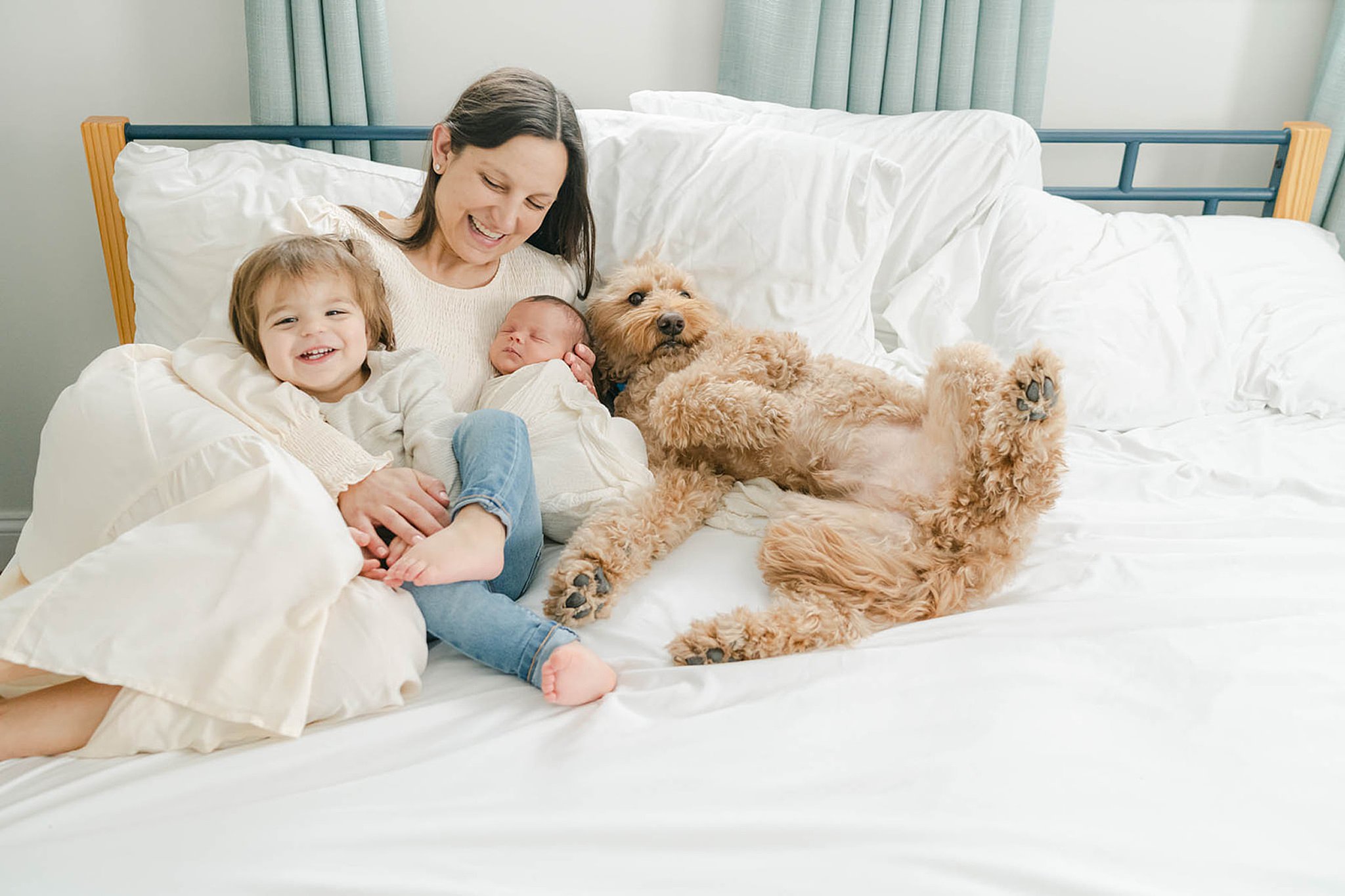 Mother, young son and newborn smile and cuddle in a white bed with their brown fluffy dog Baby Boutique West Chester PA