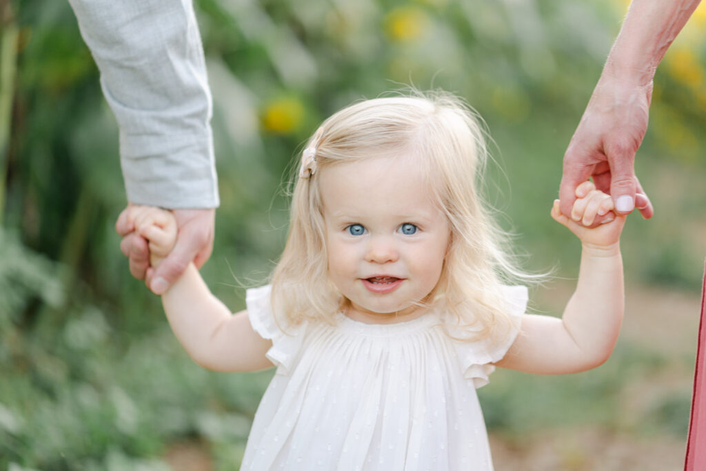 little girl with white dress and blonde hair hods parents hands smiling