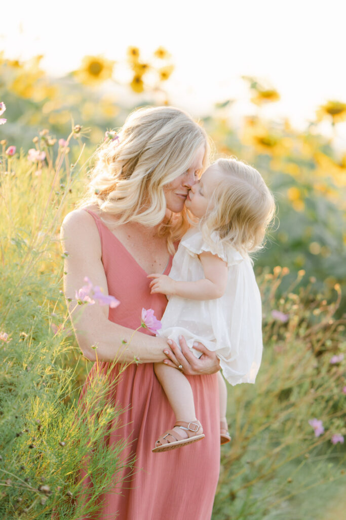 mom in long pink dress kisses baby girl in a field of sunflowers outside of Philadelphia PA