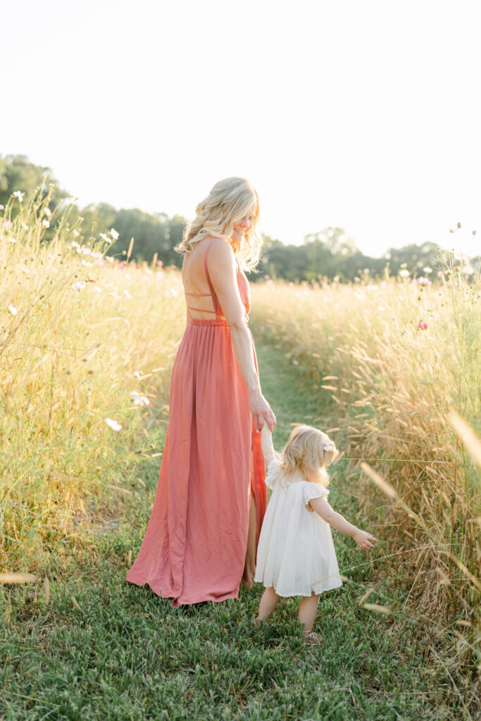 Mom in long pink dress walks through a golden field at sunset with toddler girl in white dress