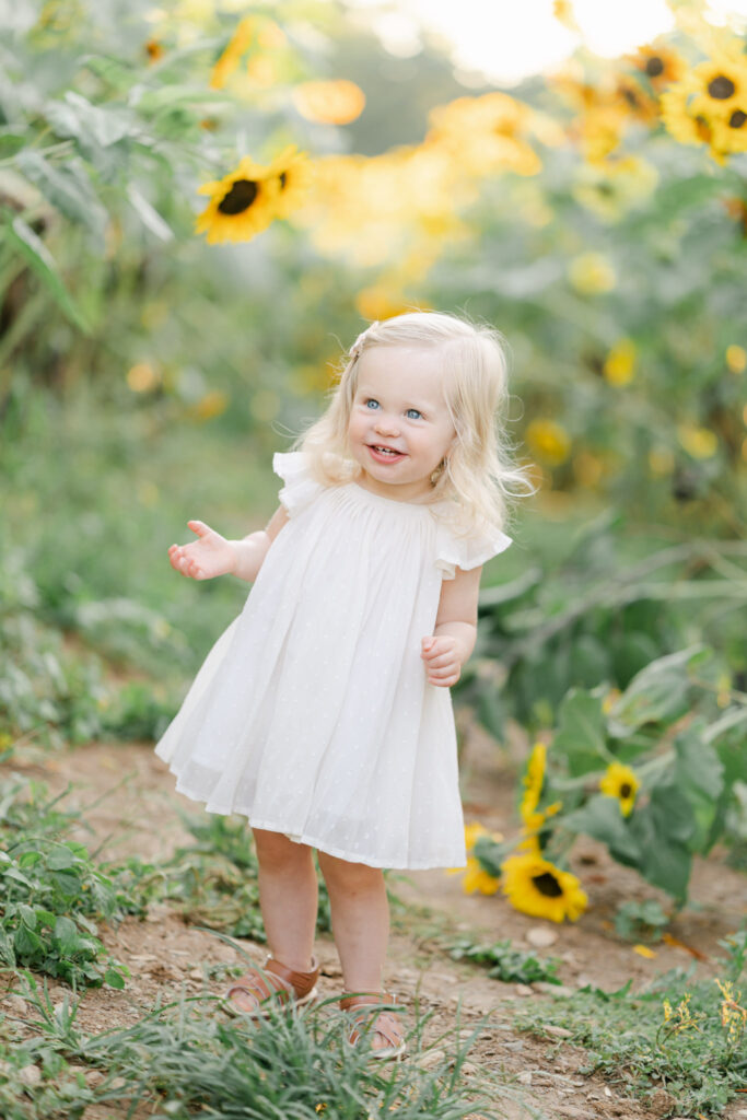 Little girl in white dress stands in a sunflower field smiling during a photo session with AnneMarie Hamant