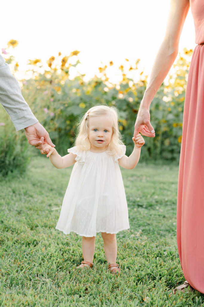 little blonde girl holds parents hands in a sunflower field outside of Philadelphia PA