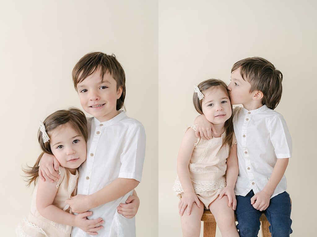 Toddler brother and sister hugging and kissing each other on the head in front of a beige backdrop